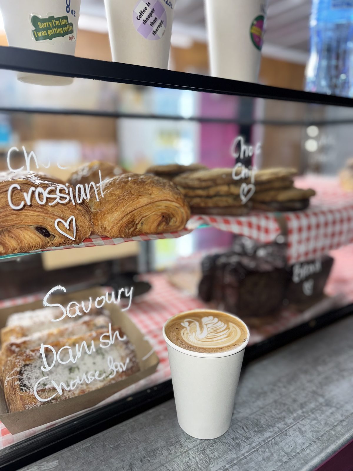 Chalk-labelled choc croissants and savoury Danish in the case with a coffee in front