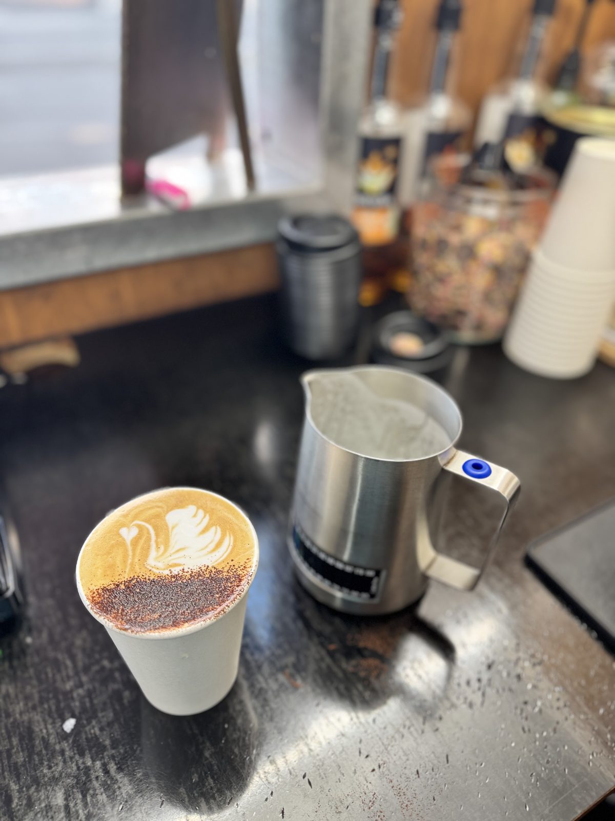 A coffee with fern latte art beside a milk jug on the counter