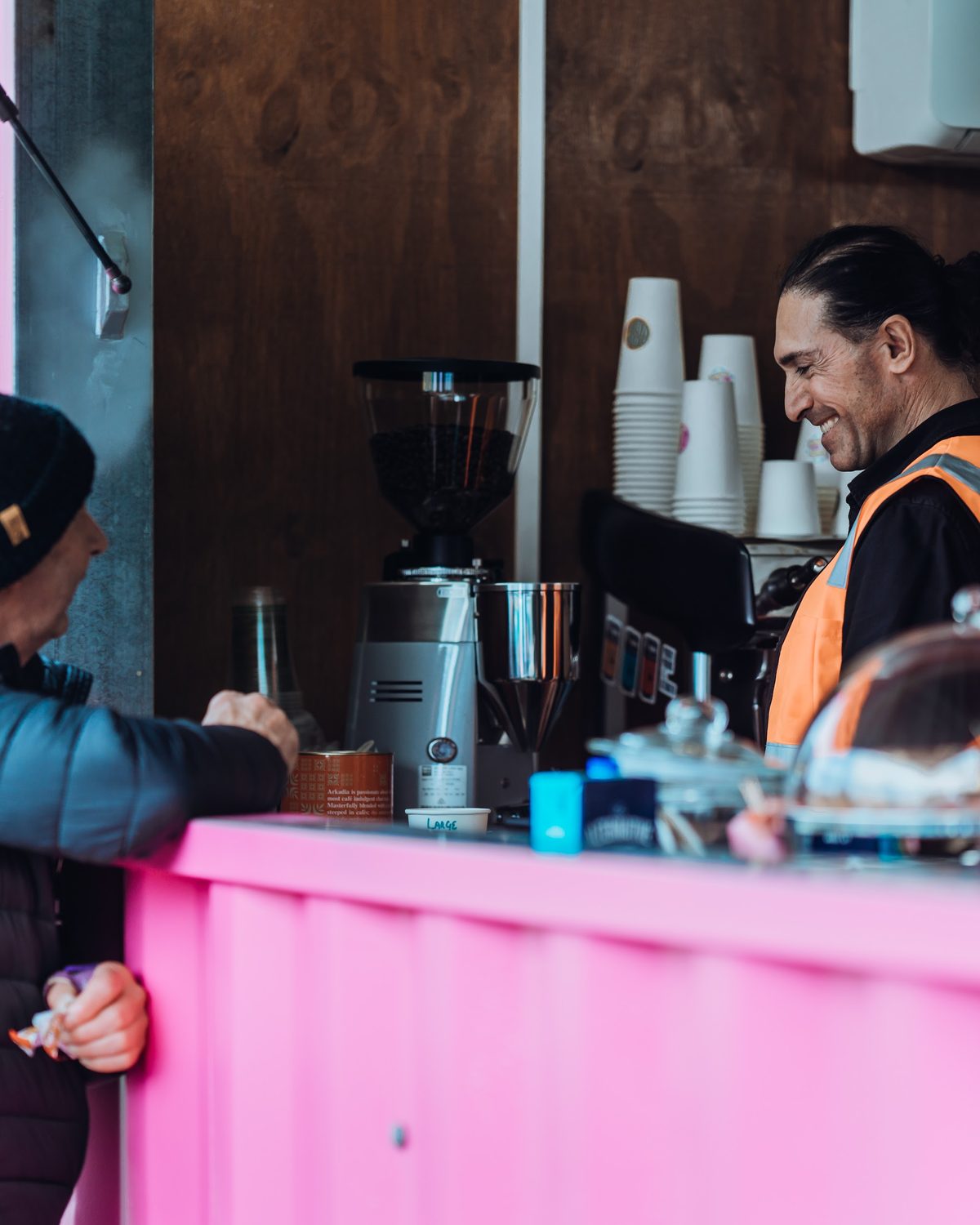 A smiling barista in hi-vis at the pink counter, espresso machine and cup stack behind